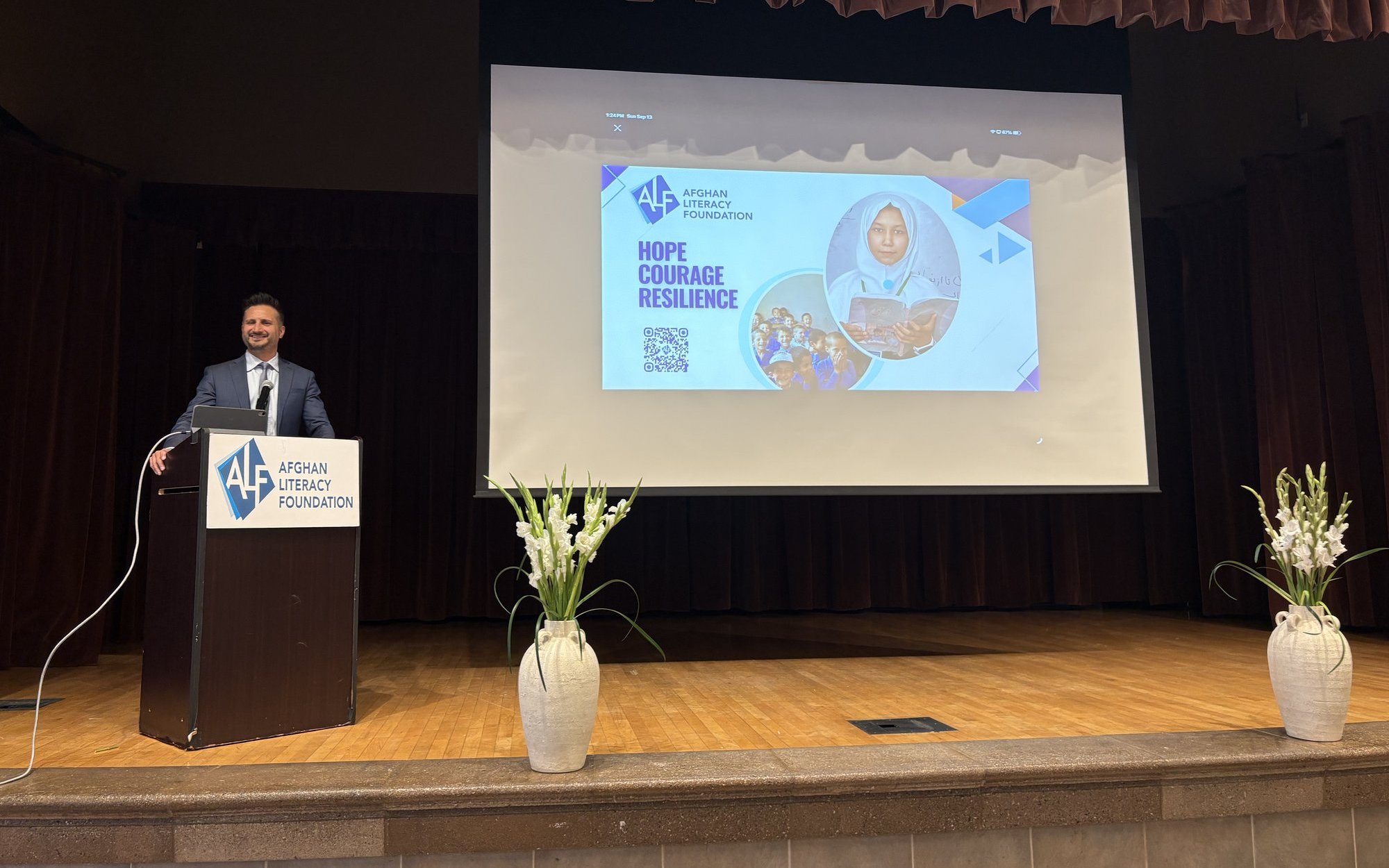 Asim Khan at the Afghan Literacy Foundation lectern on stage beside the event screen