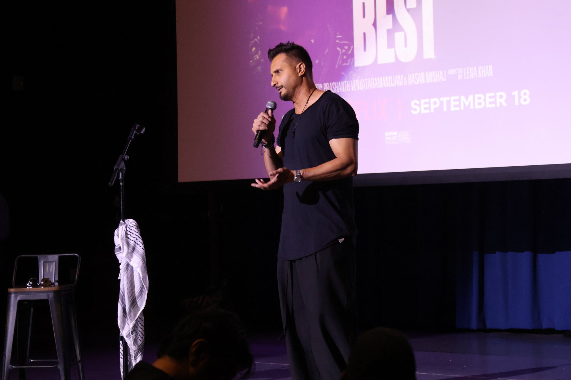 Asim Khan speaking on stage in front of the Best of the Best poster on the theater screen