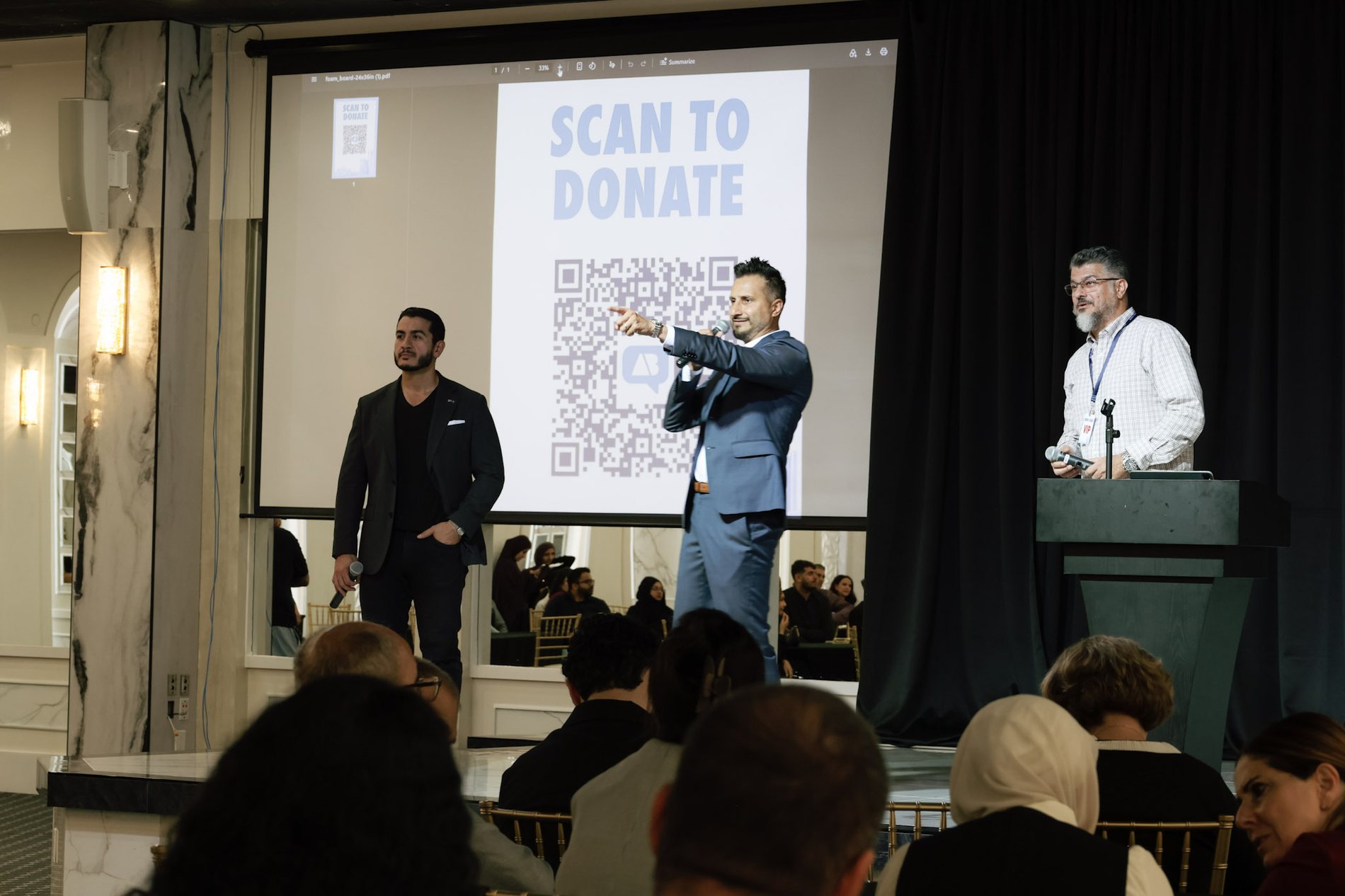 Asim Khan on stage making the ask in front of a giving screen, with Abdul El-Sayed beside him and the audience seated