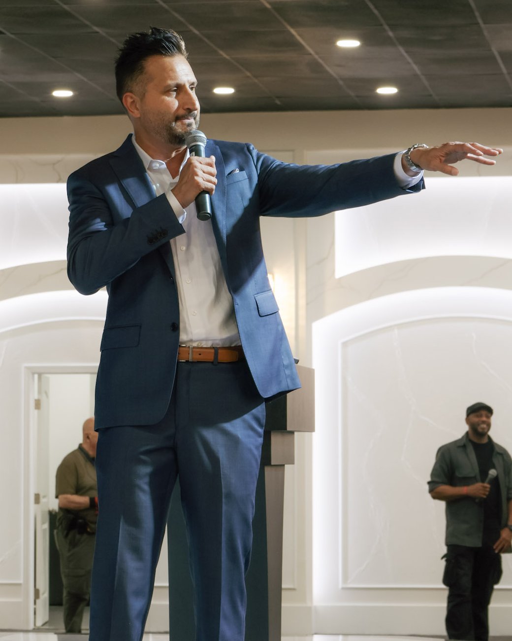 Asim Khan working the room with a handheld microphone, one arm out to the crowd